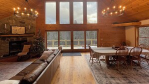 Een knus interieur van een chalet met een stenen open haard, een kerstboom, een grote eettafel met stoelen en een houten terras zichtbaar door de ramen.