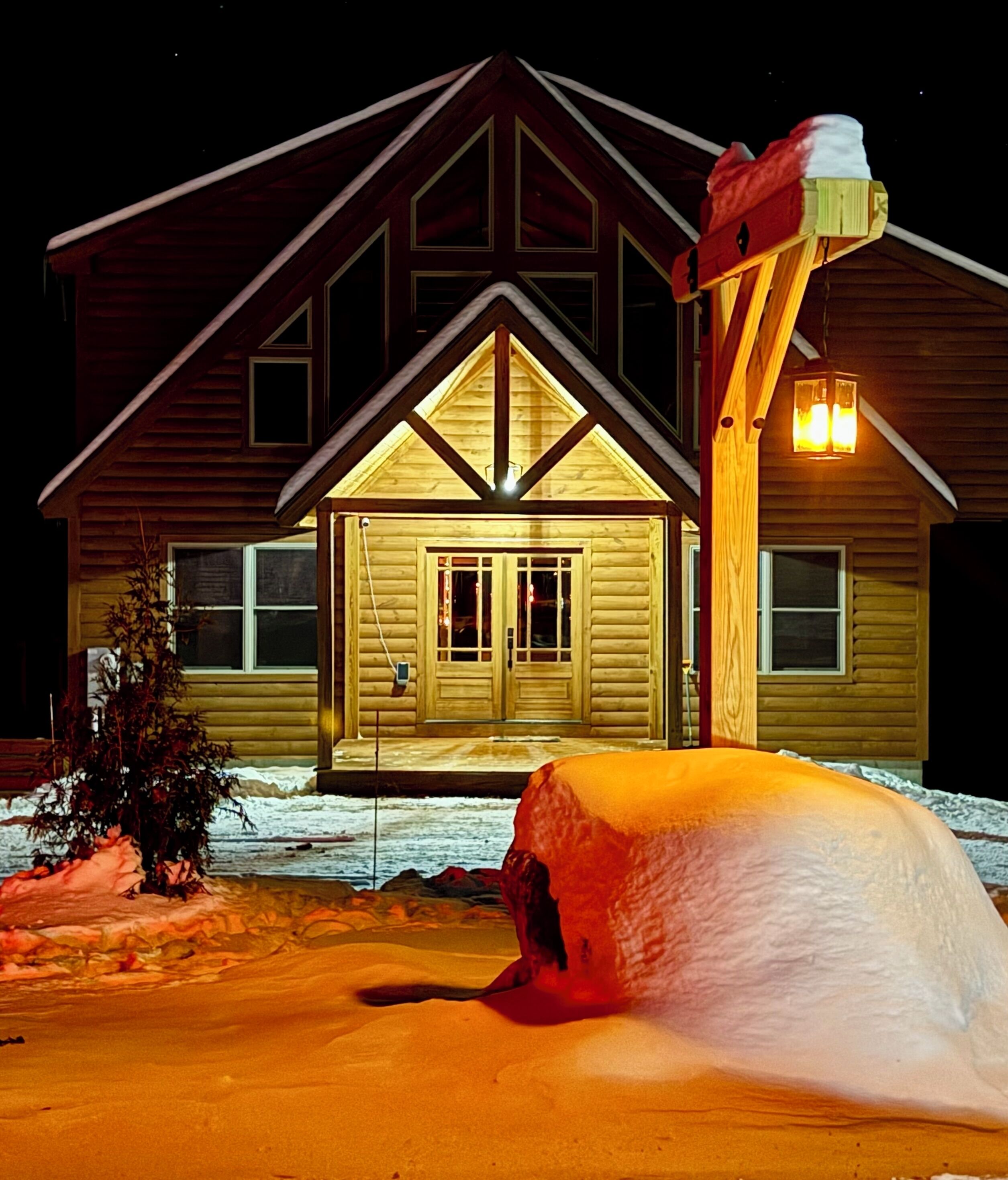 Une cabane en rondins avec une cheminée imposante, une véranda avec un rocher recouvert de neige et un amas de neige devant.