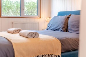 Un lit bien fait, avec des serviettes pliées, une tête de lit bleue et une fenêtre donnant sur des arbres.