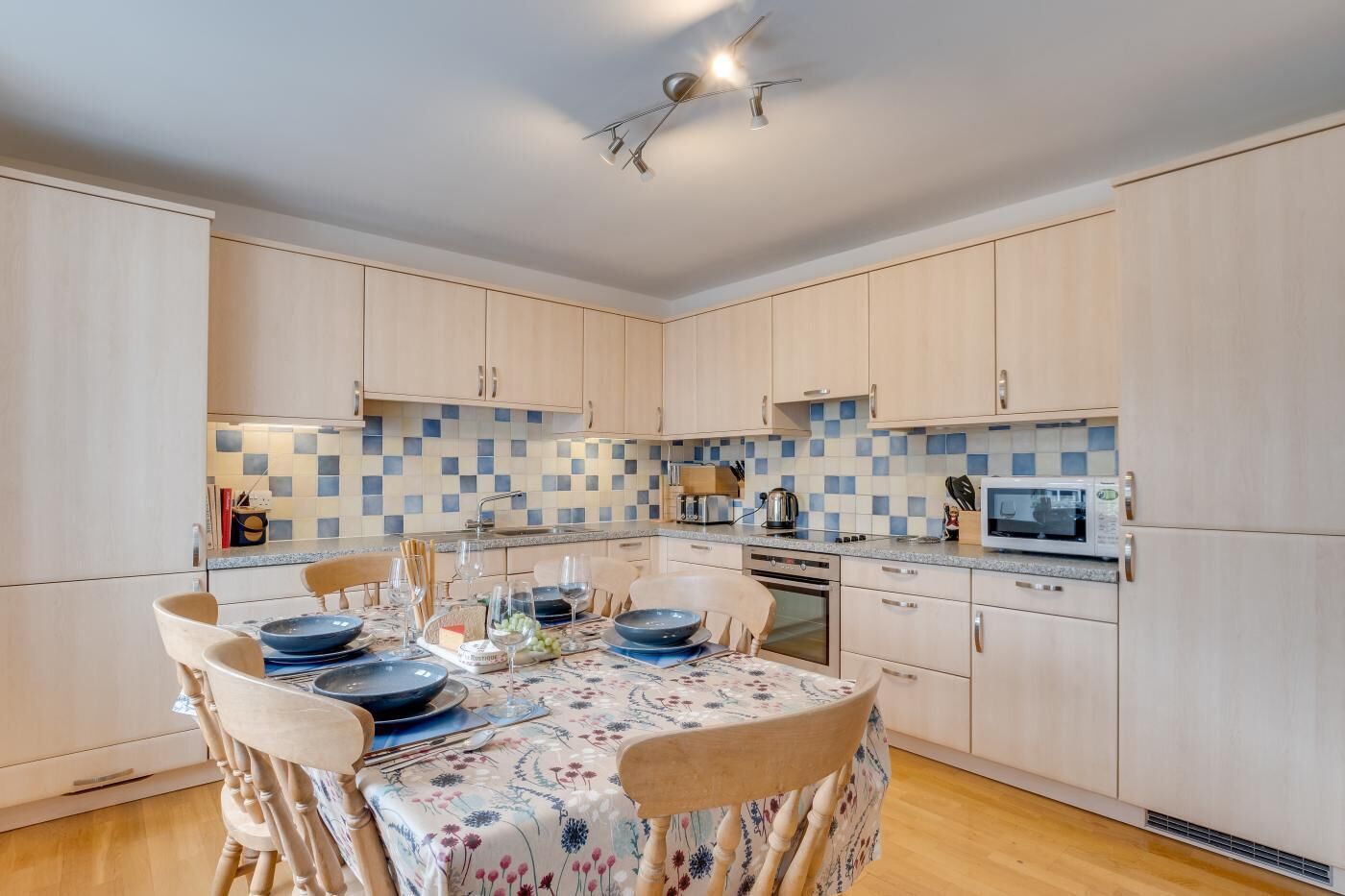 A modern kitchen with light wood cabinets, a checkered backsplash, and a dining area with a floral tablecloth.