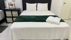Un lit bien fait, avec du linge de lit blanc et un plaid vert, une table de chevet avec une lampe et des fleurs, et une armoire en arrière-plan.