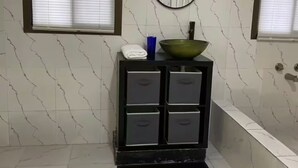 A bathroom with a marble floor, a black vanity with drawers, a green bowl sink, a round mirror, and two windows with blinds. - Lagoona Ocean Suites Monrovia Thinker’s Village (Mills Center)