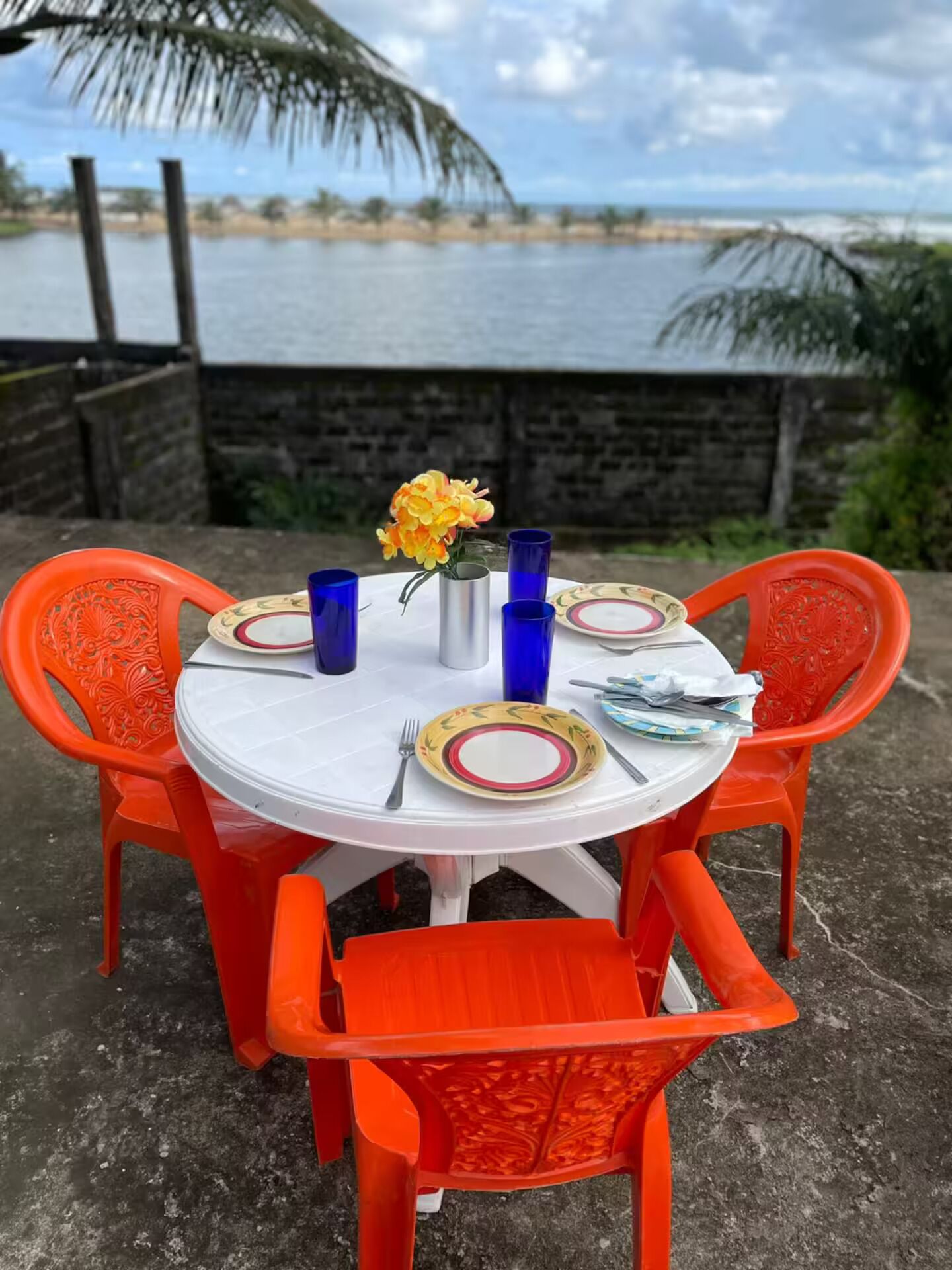 A round outdoor table with a white tablecloth, surrounded by orange chairs with intricate designs. There are blue glass tumblers, a metal pitcher, and a plate with a floral pattern. A vase with yellow flowers is placed in the center. The background features a body of water and a stone wall.