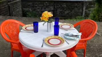 A round outdoor table with a white tablecloth, surrounded by orange chairs with intricate designs. There are blue glass tumblers, a metal pitcher, and a plate with a floral pattern. A vase with yellow flowers is placed in the center. The background features a body of water and a stone wall.