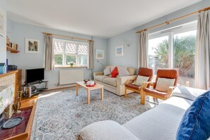 A living room with a sofa, armchairs, a coffee table, a TV, and a window with curtains. - 4 Bed in Southwold (oc-spn) (Reydon)