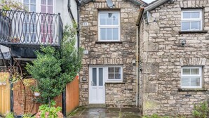 A stone building with a white door and windows, a small garden with plants, and a metal fence. - 2 Bed in Kendal (oc-gl097) (Kendal)