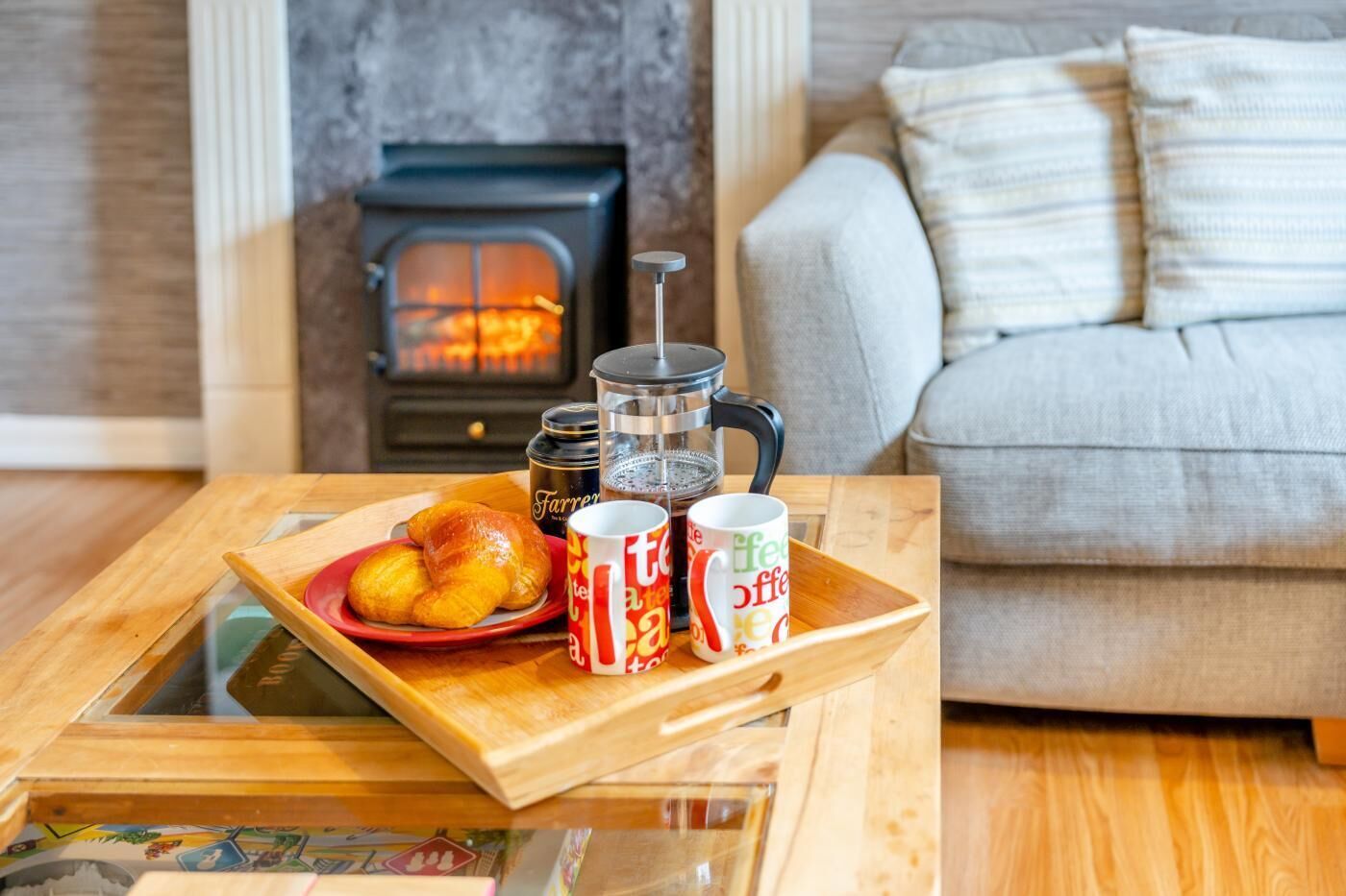 A cozy living room with a fireplace, a coffee table with croissants, a French press, and two coffee mugs.