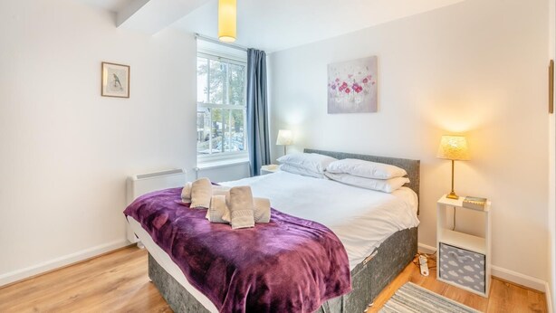 A bedroom with a bed, two bedside lamps, a window with curtains, a radiator, and a painting on the wall. - 2 Bed in Kendal (oc-gl097) (Kendal)