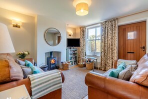 A cozy living room with leather sofas, a wood-burning stove, a television, and a bookshelf. - 2 Bed in Aldeburgh (oc-fsh) (Aldeburgh)