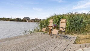 A wooden deck with two lounge chairs and a small table, overlooking a calm body of water with houses in the background. - 2 Bed in Snettisham (oc-e32982) (Snettisham)