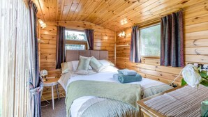 A wooden cabin bedroom with a bed, bedside table, and a window with curtains. - 2 Bed in Snettisham (oc-e32982) (Snettisham)