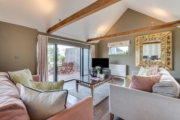 A spacious living room with a large sofa, a coffee table, and a television. There is a sliding glass door leading to an outdoor patio area with chairs and a table.