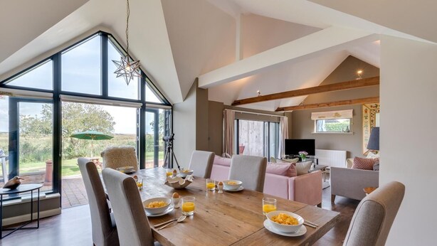 Een eetgedeelte met een houten tafel gedekt voor het ontbijt, een groot raam met uitzicht op de omgeving, en een gezellige zithoek met een televisie en zitplaatsen.