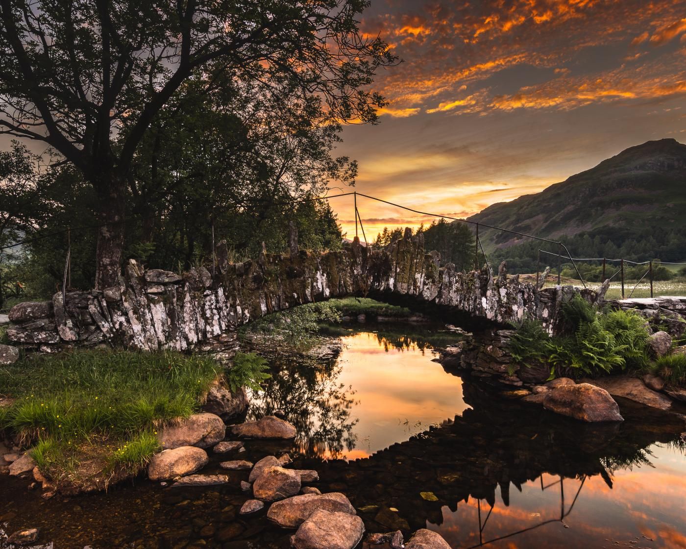A stone bridge over a calm river with a sunset in the background.