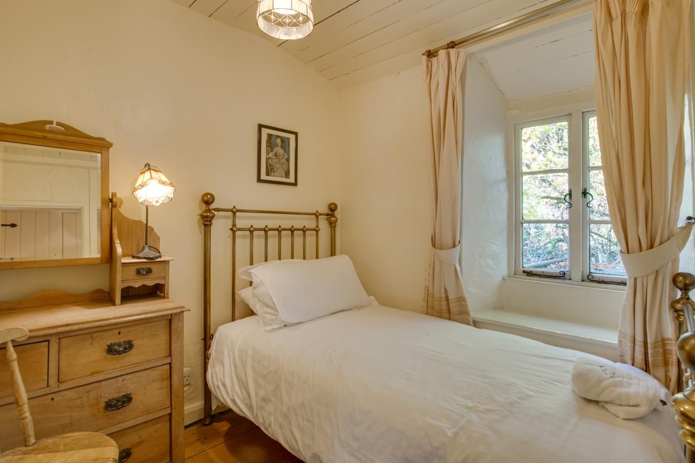 A bedroom with a bed, a bedside table, a lamp, a window with curtains, and a wall-mounted picture.