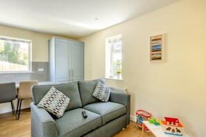 Ein Wohnzimmer mit grauem Sofa, einem kleinen Tisch mit Spielzeug und einem Fenster mit Blick auf Grünflächen.