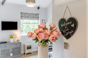 Uma sala de estar com televisão, um vaso de flores, uma placa na parede e um pequeno armário.