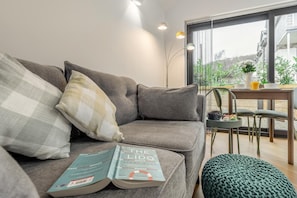 A modern living room with a grey sofa, a book titled 'The Lido,' a green knitted ottoman, and a dining area with a table and chairs. - 1 Bed in Braunton (oc-b32917) (Braunton)