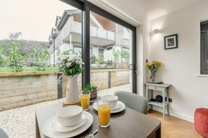 A modern room with a dining table set for breakfast, a coffee table, and a view of a wooden deck and building. - 1 Bed in Braunton (oc-b32917) (Braunton)
