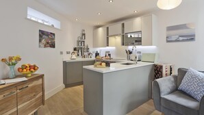 A modern kitchen with a central island, built-in appliances, and a grey sofa in the living area. - 2 Bed in Southwold (oc-wtp) (Southwold)