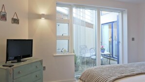 A bedroom with a TV on a dresser, a bed with a knitted blanket, a balcony with chairs, and a sliding glass door. - 2 Bed in Southwold (oc-wtp) (Southwold)