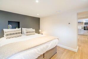Une chambre avec un lit, des tables de chevet, une commode, une étagère murale avec des livres, et un espace cuisine visible.