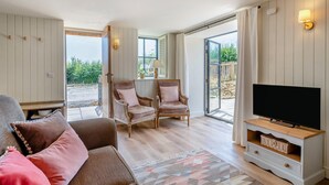 Ein gemütliches Wohnzimmer mit einer Couch, zwei Sesseln, einem Fernseher und einem großen Fenster mit Blick ins Freie.