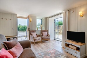 Ein gemütliches Wohnzimmer mit einer Couch, zwei Sesseln, einem Fernseher und einem großen Fenster mit Blick ins Freie.