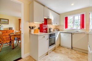 A compact kitchen with a dishwasher, oven, and a window with red curtains. - 3 Bed in Thorpeness (oc-ab1d) (Thorpeness)
