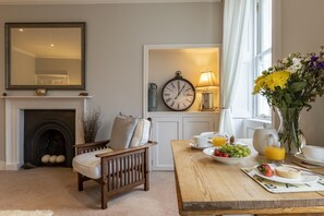 A cozy living room with a fireplace, a dining table set with breakfast, a clock, and a vase of flowers. - 1 Bed in Bath (oc-bhr12) (Bath)