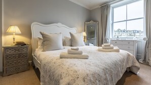 Een keurig opgemaakt slaapkamer met een bed, nachtkastjes, een lamp en een uitzicht op de stad door een raam.