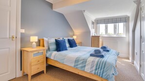 Un dormitorio con techo inclinado, una cama con ropa de cama azul, una mesita de noche con lámpara y una ventana con persianas.