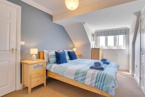 Ein Schlafzimmer mit schrägen Wänden, einem Bett mit blauer Bettwäsche, einem Nachttisch mit Lampe und einem Fenster mit Jalousien.