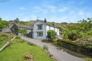 A white two-story house with a stone exterior, surrounded by lush greenery and a gravel path leading to the entrance. - 3 Bed in Elterwater (oc-gl011) (Chapel Stile)