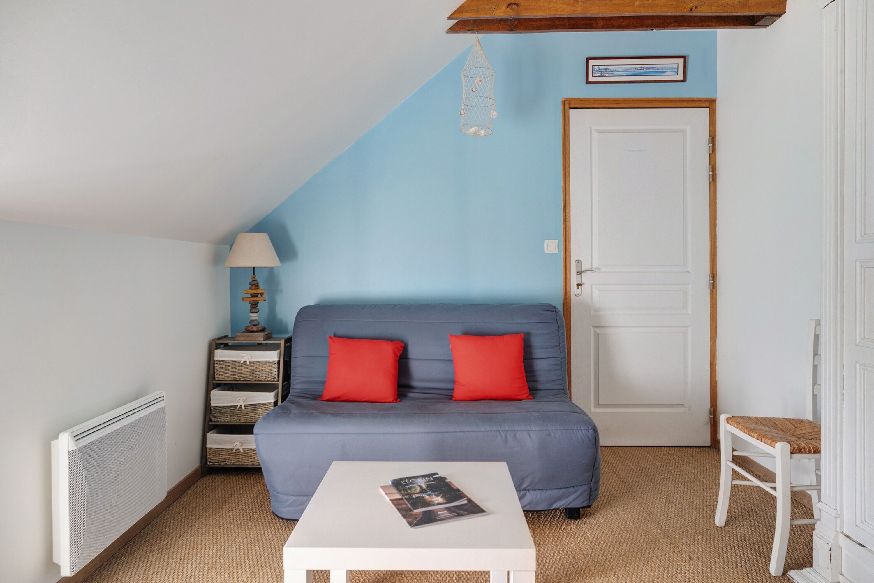 Een kamer met een blauwe bank, een witte tafel, een rieten stoel, een lamp en een deur.