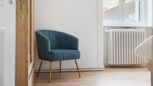 Een kamer met een blauwgroene fauteuil, houten vloer, witte wanden en een raam met een radiator.