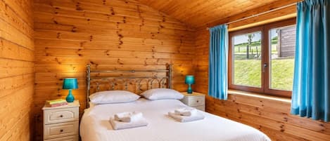 Quarto de cabana de madeira com cama, duas mesas de cabeceira e janela com cortinas.