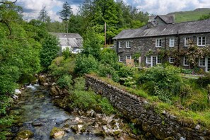 A stone wall beside a flowing stream, surrounded by lush greenery and a stone building with multiple windows. - 2 Bed in Elterwater (oc-gl053) (Chapel Stile)