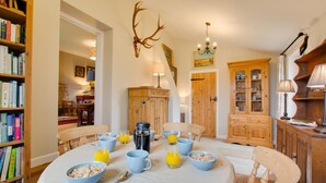Ein Essbereich mit einem runden Tisch, der für das Frühstück gedeckt ist, umgeben von Holzstühlen. Eine Bücherwand mit Büchern befindet sich links, und ein Holzschrank mit Glastüren rechts.