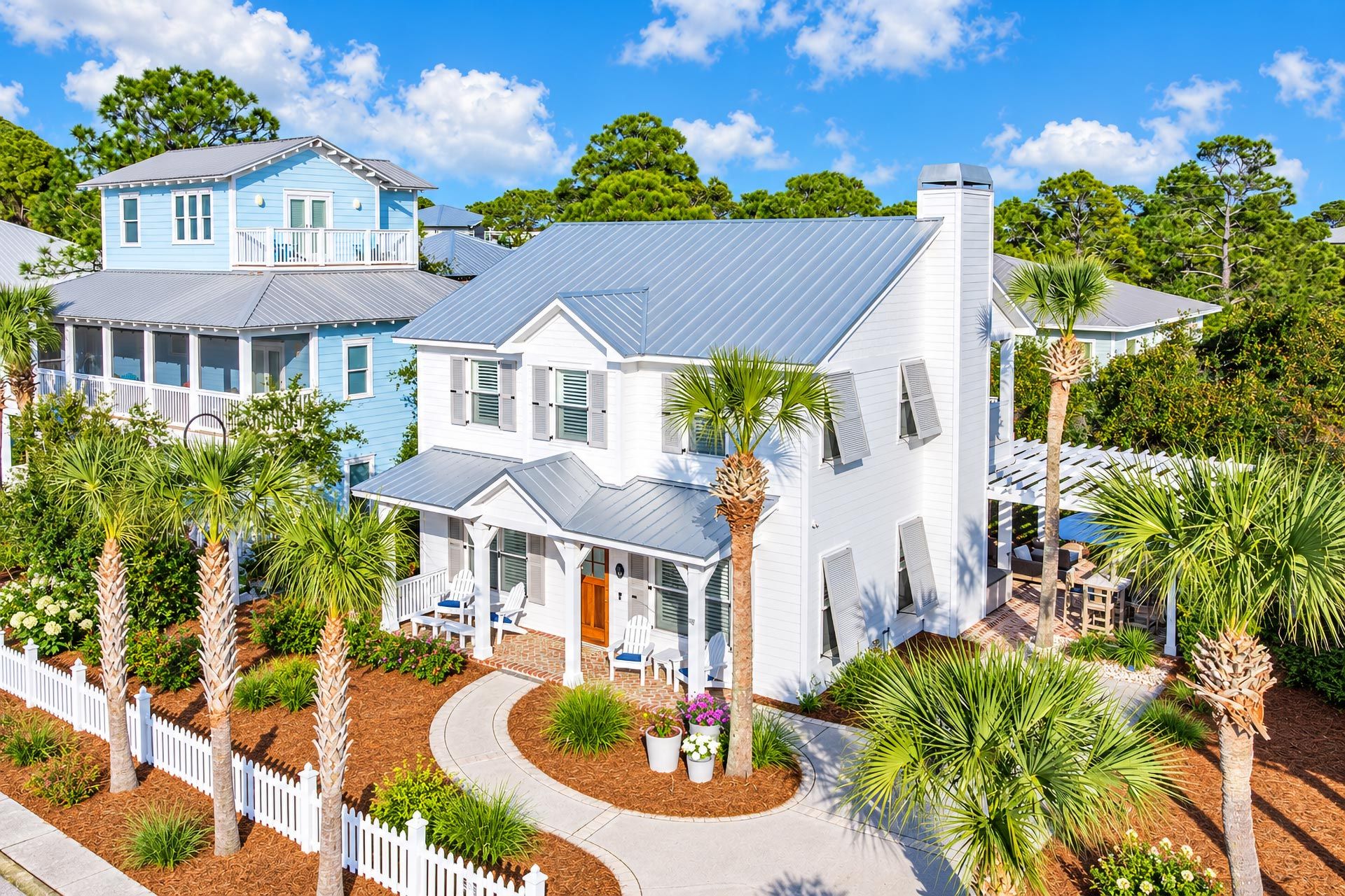 A coastal house with a covered porch, a white fence, and a variety of tropical plants.