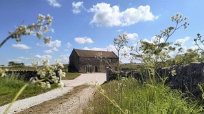 Un bâtiment en pierre, accessible par un chemin de gravier, entouré de verdure et sous un ciel dégagé.
