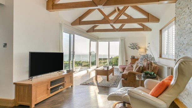 A spacious living room with a large TV, wooden furniture, a comfortable sofa, and a view of the outdoors. - 3 Bed in Newlyn (oc-s33015) (Newlyn)
