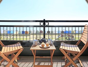 A balcony with two wooden chairs, a table with a teapot, cups, and a plate with food, overlooking a beach with colorful beach huts and a playground. - 3 Bed in Felixstowe (oc-fel) (Felixstowe)