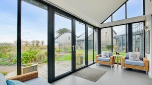 Una habitació àmplia amb grans portes de vidre que donen a un jardí, mobles de vimet i vistes cap a una casa.