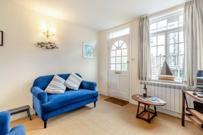 Ein Wohnzimmer mit einem blauen Sofa, einem kleinen Tisch mit Flasche und Gläsern, einem Fernseher und einem Fenster mit Vorhängen.