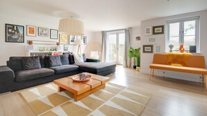 Ein modernes Wohnzimmer mit einem grauen Ecksofa, einem Holztisch und einer gelben Bank. An den Wänden befinden sich gerahmte Bilder, ein großes Fenster mit Vorhängen und ein Blumentopf.