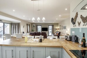 A modern kitchen with a central island, a dining area with a table and chairs, a living room with a sofa and a TV, and a kitchen counter with appliances and a bottle of champagne. - 2 Bed in Horning (oc-h33013) (Horning)