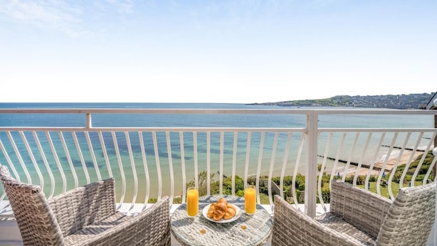 Varanda com cadeiras de vime, uma mesa com croissants e suco, e vista para o mar e terra ao longe.