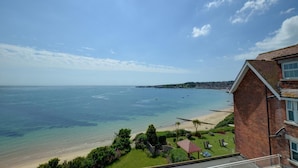 Vista costeira com céu claro, praia de areia e um edifício residencial com telhado inclinado.