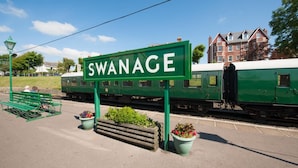 Uma placa verde com a inscrição 'SWANAGE', um banco, um trem e um edifício ao fundo.